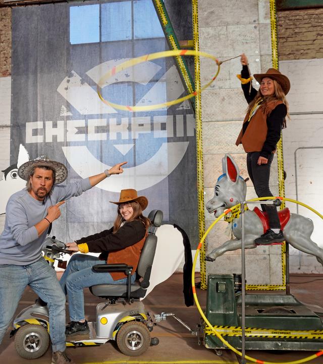 Checkpoint: Tommy Scheel zeigt auf Maja, die Lassoschwingend auf einem Karussell-Esel steht, der von einem Senioren-Scooter gezogen wird, der von Lisa gefahren wird. 