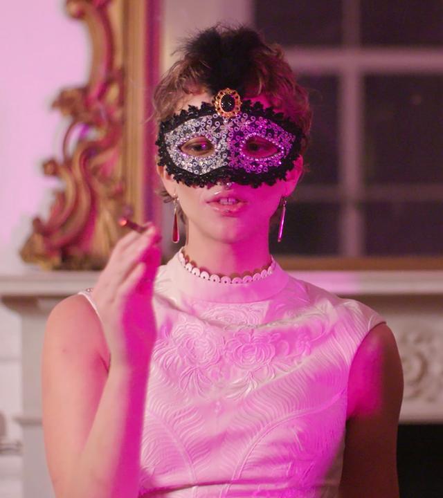 Eine kurzhaarige Frau in weißem Abendkleid und mit einer auffällig verzierten Augenmaske sitzt vor einem offenen Kamin. In der einen Hand hält sie eine Zigarette.