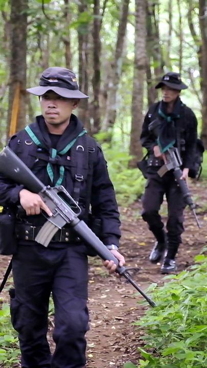 Drei Männer mit Hüten und schwarzen Uniformen laufen hintereinander über einen Waldweg. Alle drei halten schwere Maschinengewehre in den Händen.