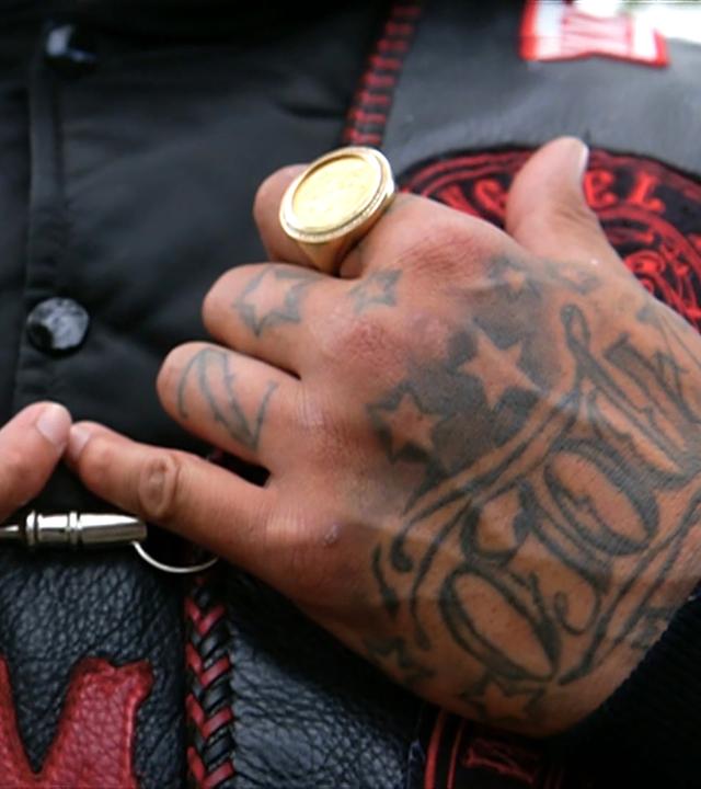 Zwei tätowierte Hände mit großen Goldringen ruhen auf der Brust einer Person, die eine Biker-Lederweste trägt.