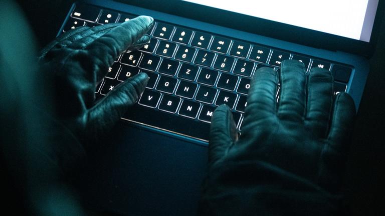 Eine Person tippt mit behandschuhten Händen auf einer beleuchteten Laptop-Tastatur. (Gestellte Szene)