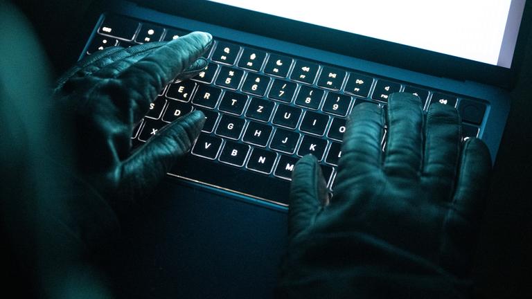 Eine Person tippt mit behandschuhten Händen auf einer beleuchteten Laptop-Tastatur. 