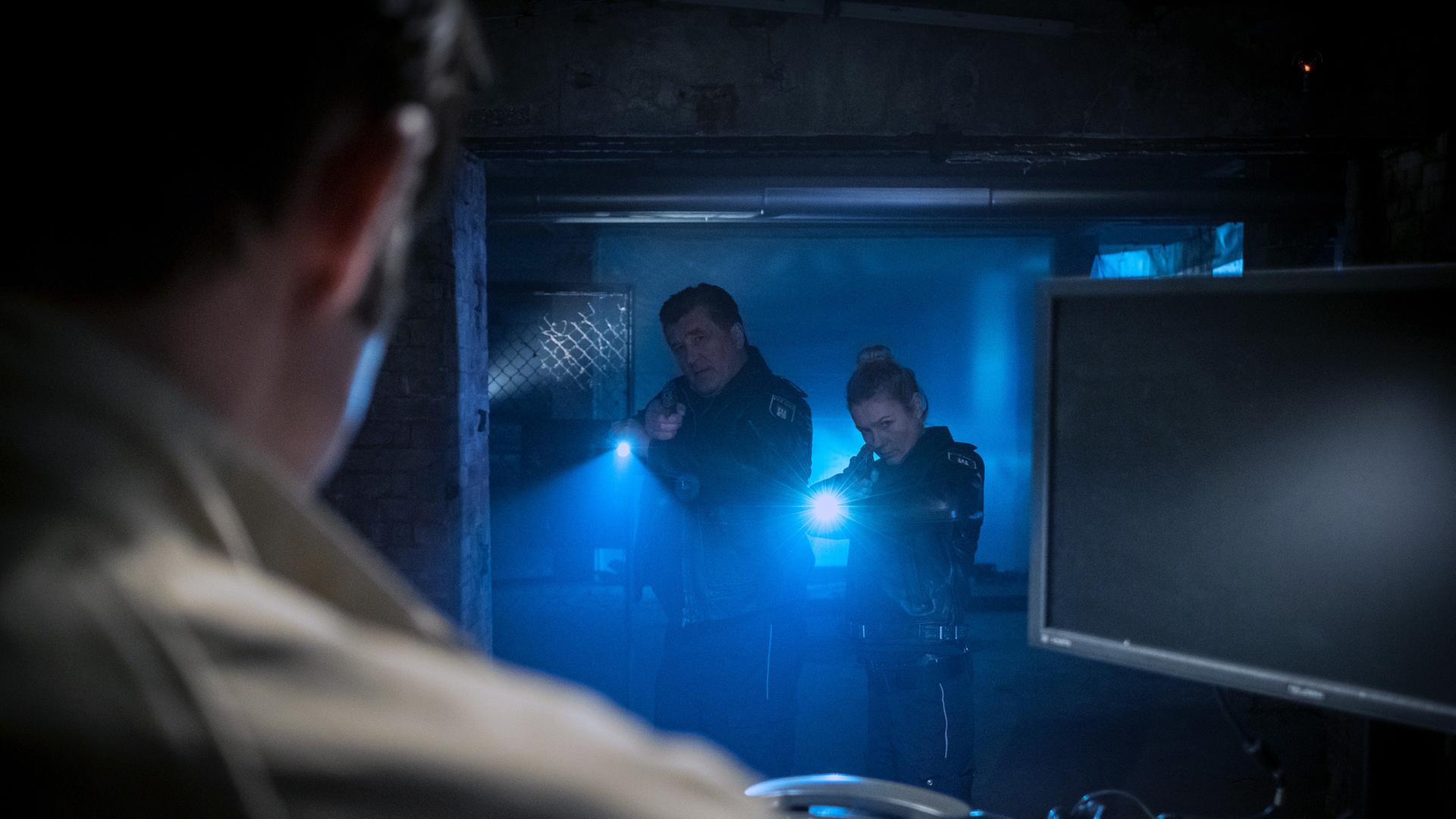 "Notruf Hafenkante - Cybermobbing": Die Polizisten Hans Moor (Bruno F. Apitz) und Franzi Jung (Rhea Harder-Vennewald) stehen nebeneinander in einem dunklen Kellerdurchgang. Beide zielen mit ihrer Waffe auf einen Mann (Paul Walther), der vor ihnen an einem Schreibtisch sitzt.