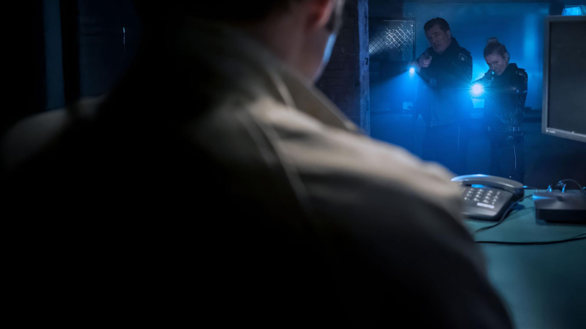 "Notruf Hafenkante - Cybermobbing": Die Polizisten Hans Moor (Bruno F. Apitz) und Franzi Jung (Rhea Harder-Vennewald) stehen nebeneinander in einem dunklen Kellerdurchgang. Beide zielen mit ihrer Waffe auf einen Mann (Paul Walther), der vor ihnen an einem Schreibtisch sitzt.