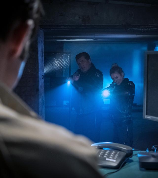 "Notruf Hafenkante - Cybermobbing": Die Polizisten Hans Moor (Bruno F. Apitz) und Franzi Jung (Rhea Harder-Vennewald) stehen nebeneinander in einem dunklen Kellerdurchgang. Beide zielen mit ihrer Waffe auf einen Mann (Paul Walther), der vor ihnen an einem Schreibtisch sitzt.