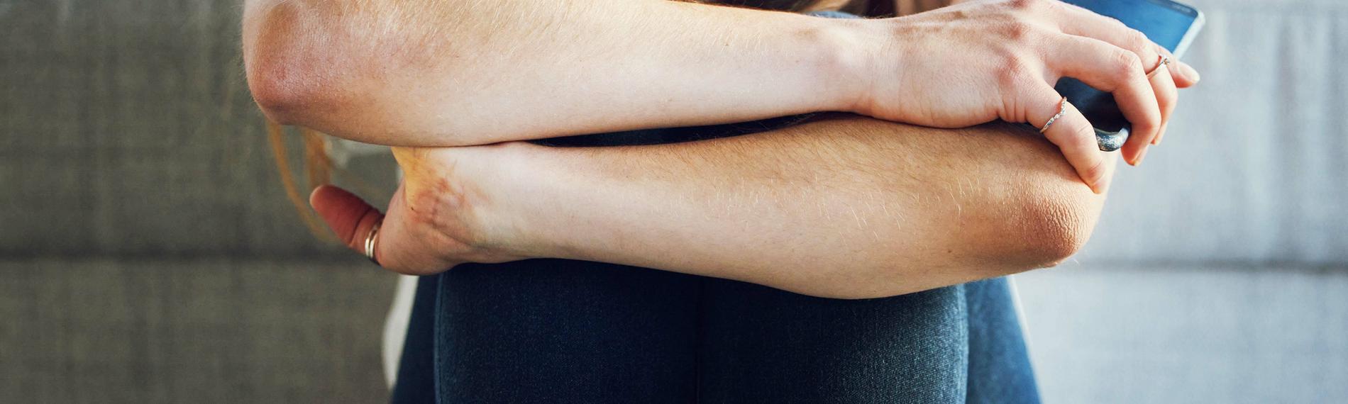 Symbolbild: Traurige junge Frau mit Smartphone kauert vor Sofa