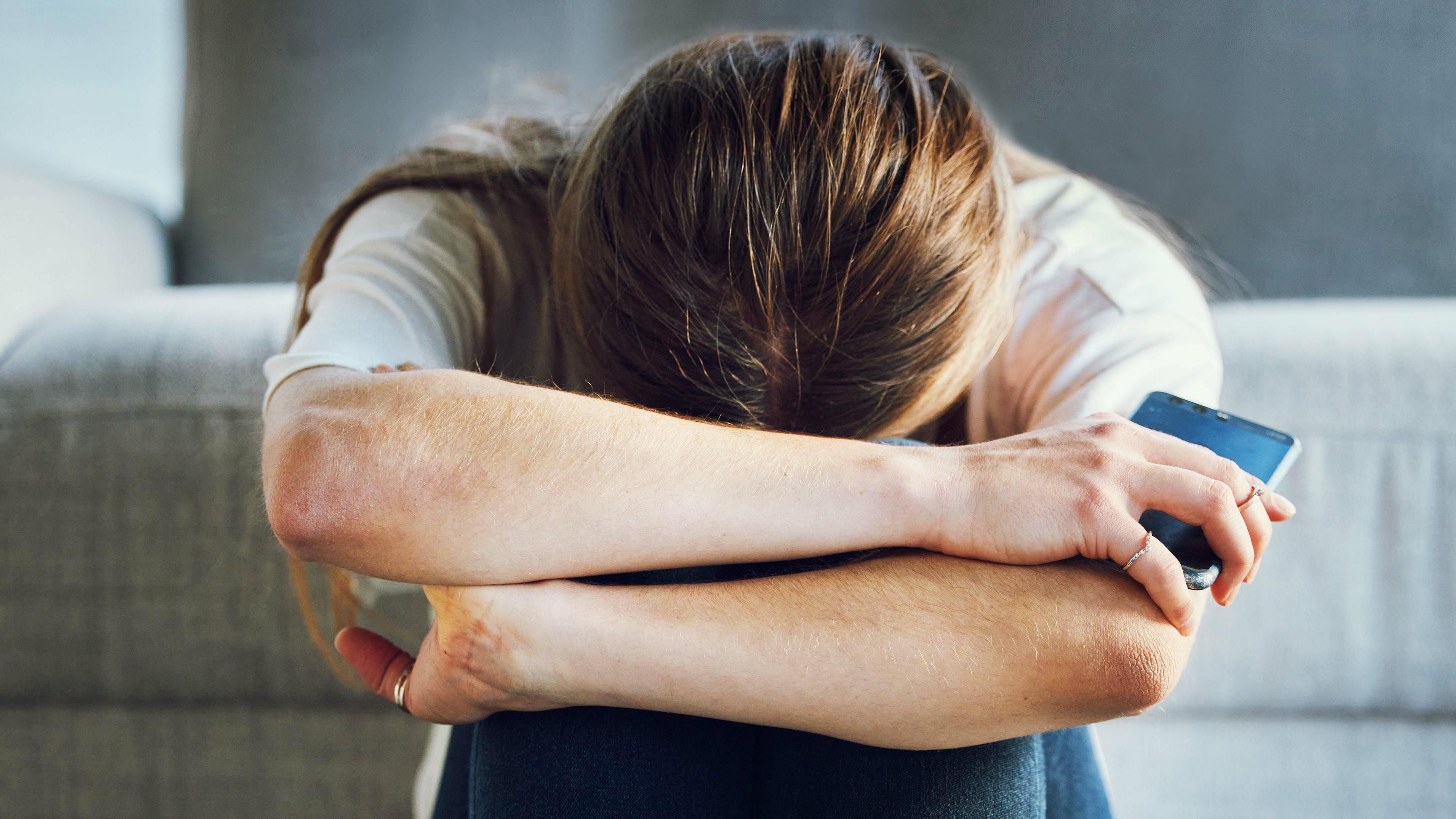 Symbolbild: Traurige junge Frau mit Smartphone kauert vor Sofa