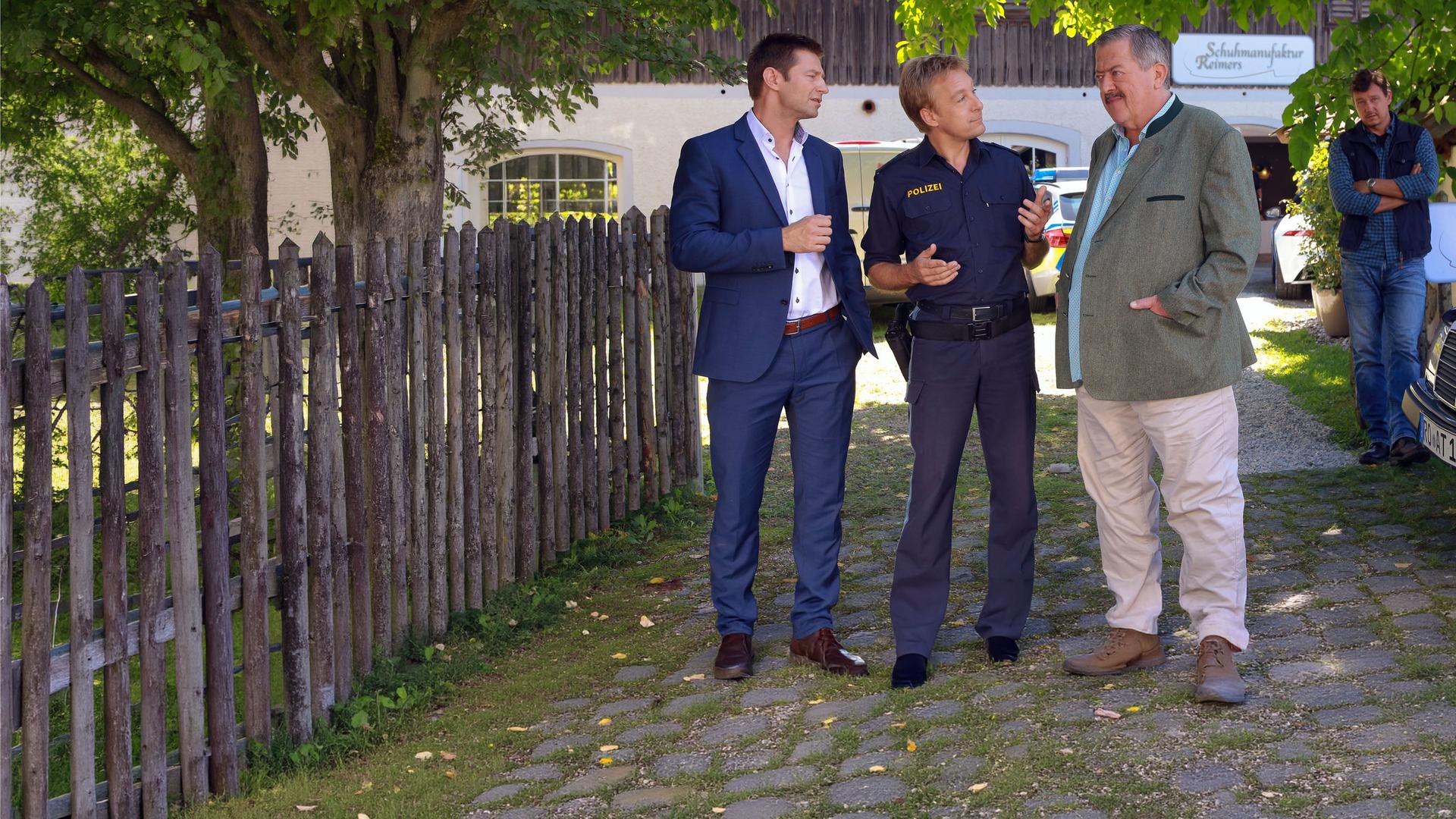 "Die Rosenheim-Cops - Da drückt der Schuh": Kommissar Hansen (Igor Jeftic), Polizist Mohr (Max Müller) und Kommissar Hofer (Joseph Hannesschläger) stehen in der Auffahrt zu einem Haus mit der Aufschrift "Schuhmanufaktur Reimers". Seitlich am Gartenzaun, an dem das gleiche Schild prangt, steht Maik Duschinsky (Markus Langer) mit verschränkten Armen. Er betrachtet die Ermittler.