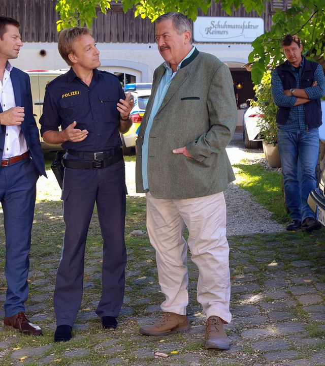 "Die Rosenheim-Cops - Da drückt der Schuh": Kommissar Hansen (Igor Jeftic), Polizist Mohr (Max Müller) und Kommissar Hofer (Joseph Hannesschläger) stehen in der Auffahrt zu einem Haus mit der Aufschrift "Schuhmanufaktur Reimers". Seitlich am Gartenzaun, an dem das gleiche Schild prangt, steht Maik Duschinsky (Markus Langer) mit verschränkten Armen. Er betrachtet die Ermittler.