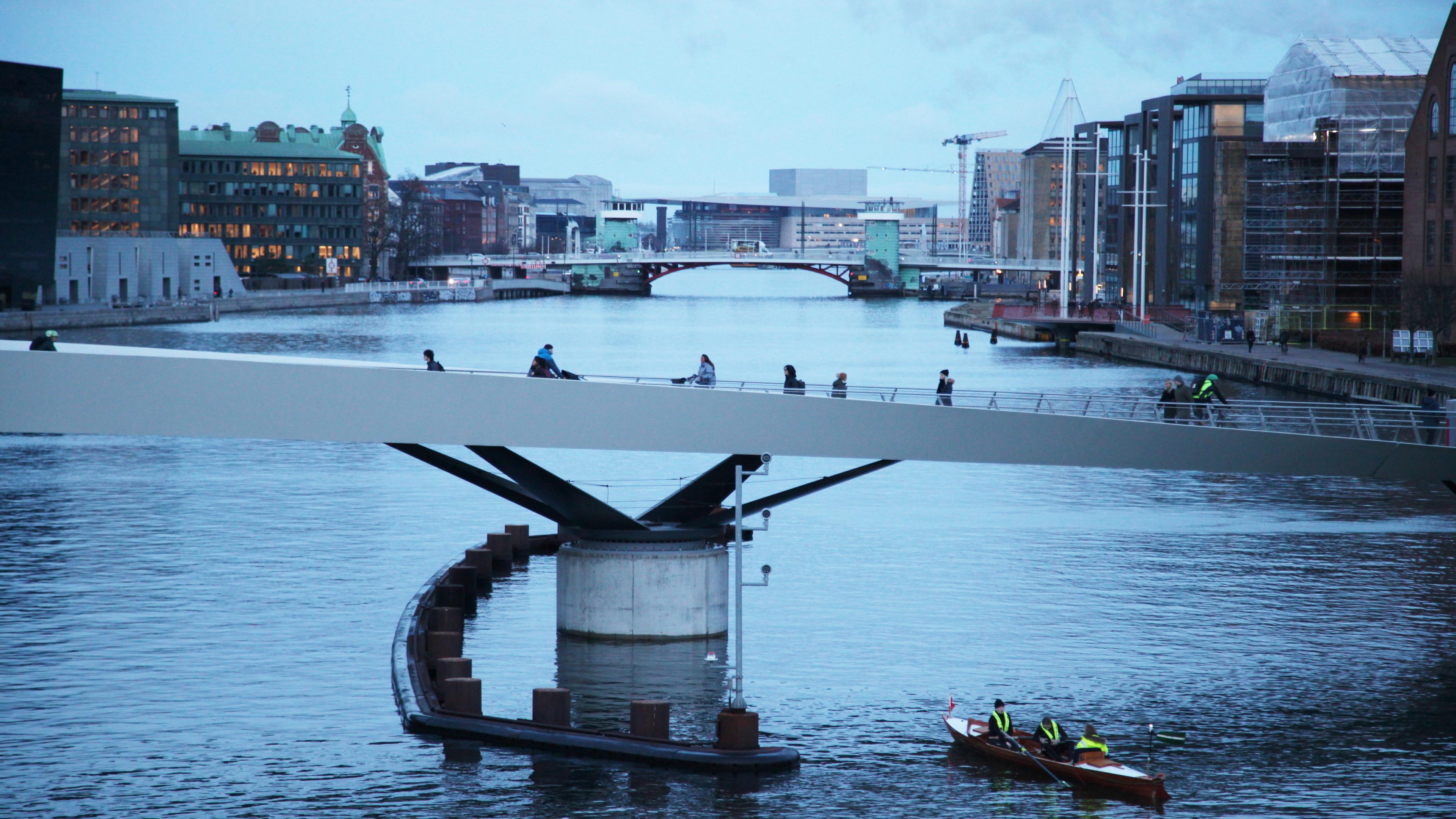 Dänemark, Kopenhagen: Radfahrer fahren über die Lille Langebro im Zentrum von Kopenhagen, während ein Ruderboot unter der Brücke hindurchfährt.