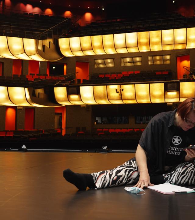 Tänzer schaut sich vor der Probe nochmal die Choreografie an. Er sitzt auf einer dunklen Bühne , während er mit dem Handy in der Hand und Paieren auf dem Boden sich die Schritte einprägt.