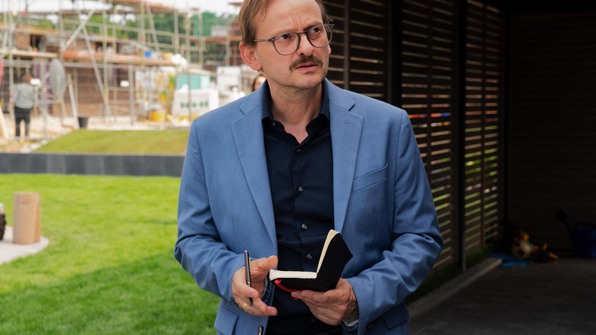 Vor dem Haus der Bressins. Danowski (Milan Peschel) hält sein Notizbuch in der Hand und blickt misstrauisch drein.