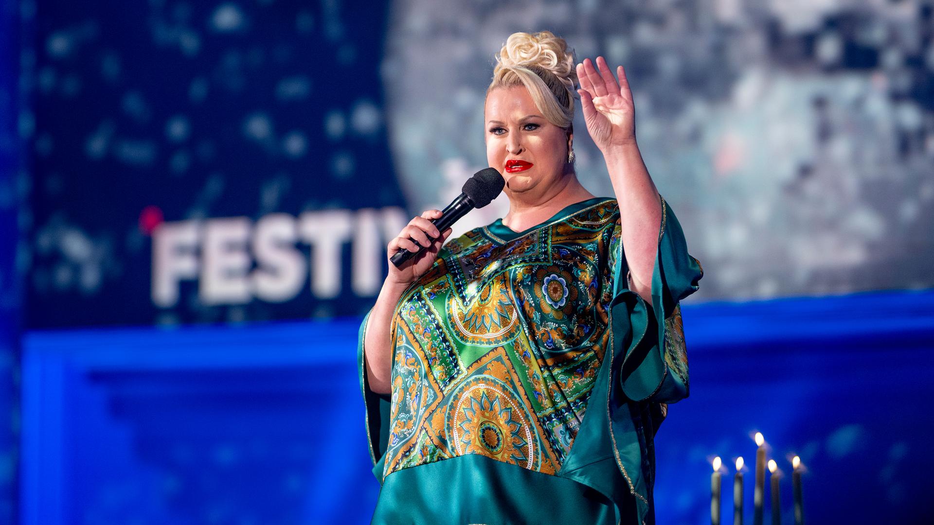 Daphne Deluxe steht in der Mitte des Bildes. Sie trägt ein blaugrünes Oberteil und hebt ihre linke Hand. Der Hintergrund ist blau beleuchtet.