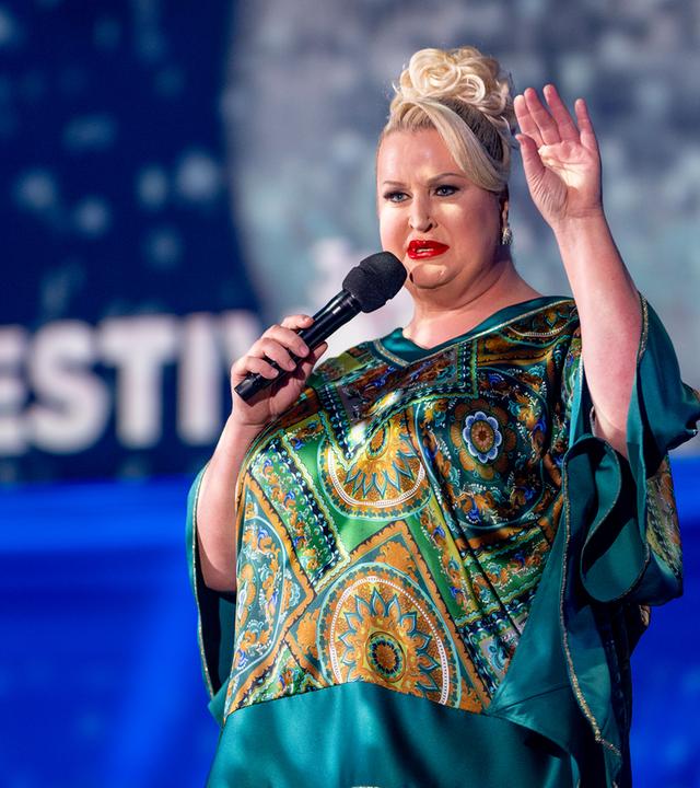 Daphne Deluxe steht in der Mitte des Bildes. Sie trägt ein blaugrünes Oberteil und hebt ihre linke Hand. Der Hintergrund ist blau beleuchtet.