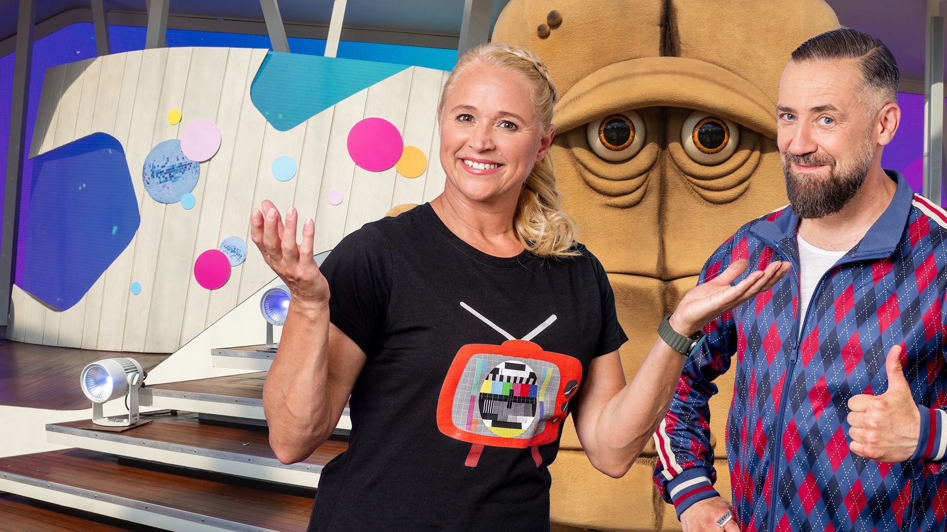 Moderator Bürger Lars Dietrich und Moderatorin Singa Gätgens stehen gut gelaunt in einem Studio. In der Mitte steht Bernd, das Brot und schaut schlecht gelaunt aus. 