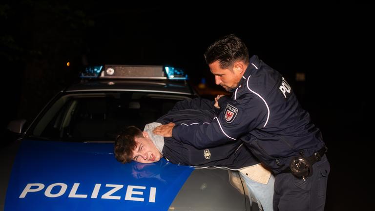 Ein Junge wird von einem Polizisten auf die Motorhaube eines Polizeiautos gedrückt.