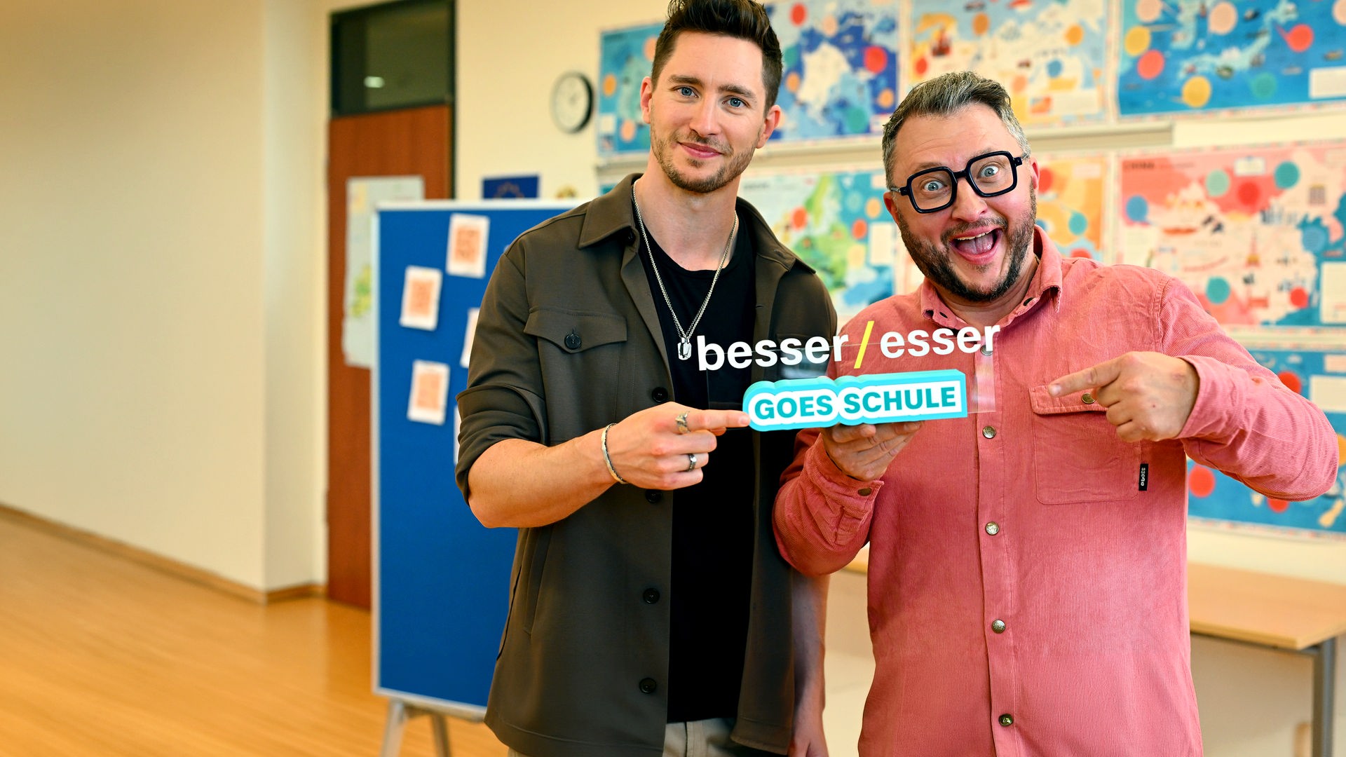 "besseresser Kids": Sebastian Lege steht rechts neben Doc Felix in einer in einem Klassenzimmer. Im Hintergrund sieht man Poster an der Wand. Gemeinsam halten sie das Logo "besseresser goes Schule" in der Hand.