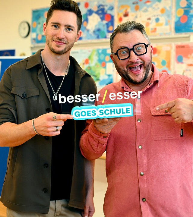 "besseresser Kids": Sebastian Lege steht rechts neben Doc Felix in einer in einem Klassenzimmer. Im Hintergrund sieht man Poster an der Wand. Gemeinsam halten sie das Logo "besseresser goes Schule" in der Hand.