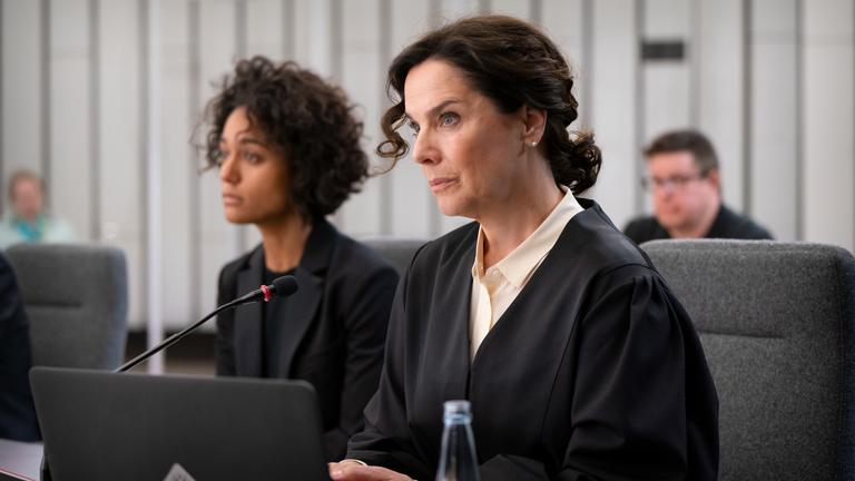 Anwältin Anna Conti (Désirée Nosbusch) sitzt in Anwaltsrobe im Gerichtssaal. Neben ihr hat ihre Mandantin und des Mordes angeklagte Sängerin Liz Jordan (Larissa Sirah Herden) Platz genommen.