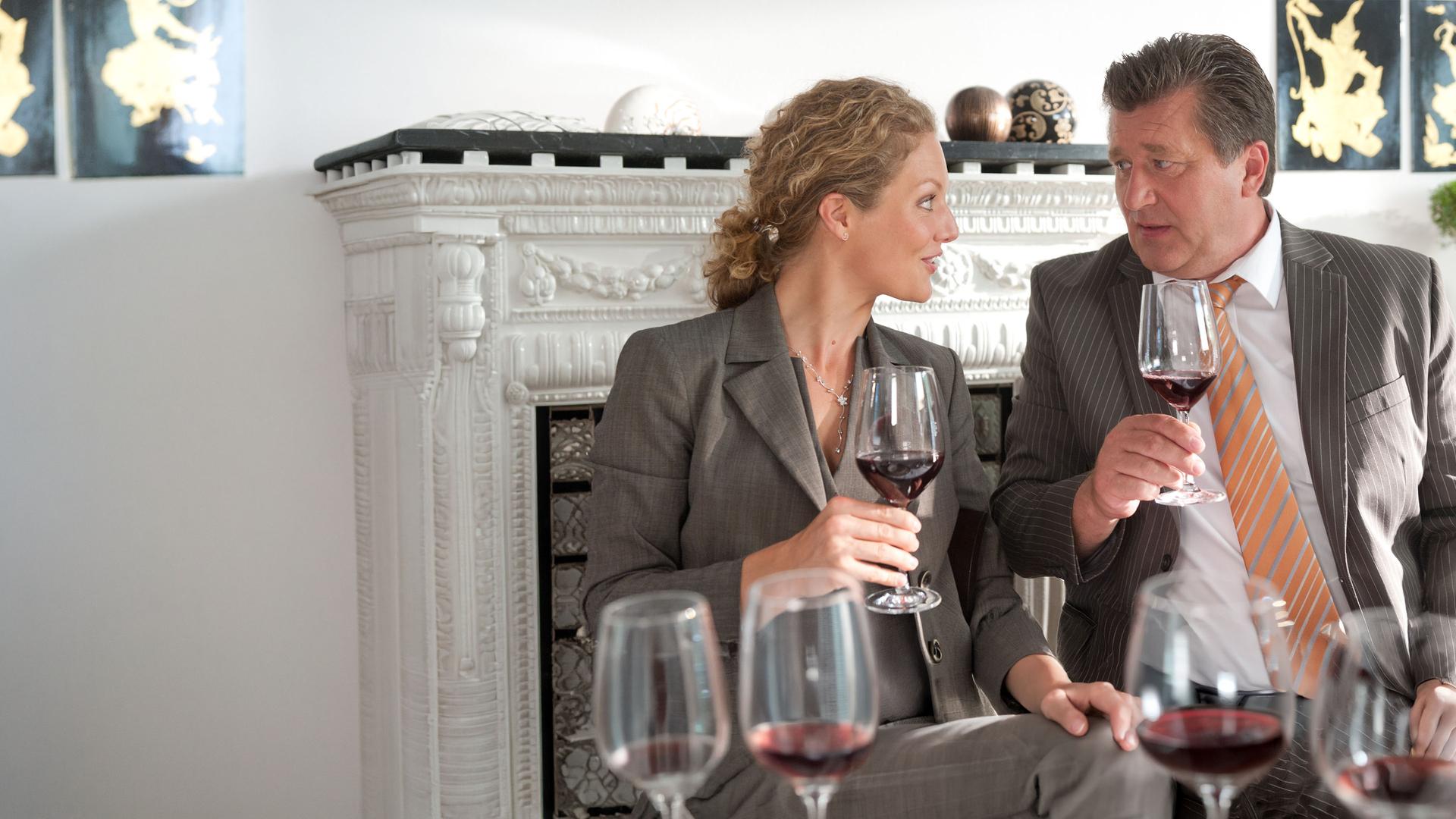 "Notruf Hafenkante - Das 1 x 1 des guten Tons": Hans (Bruno F. Apitz), der Zivilkleidung trägt und Sonja Tarrach (Tessa Mittelstaedt) sitzen nebeneinander, halten je ein Glas Rotwein in der Hand und sehen sich in die Augen.
