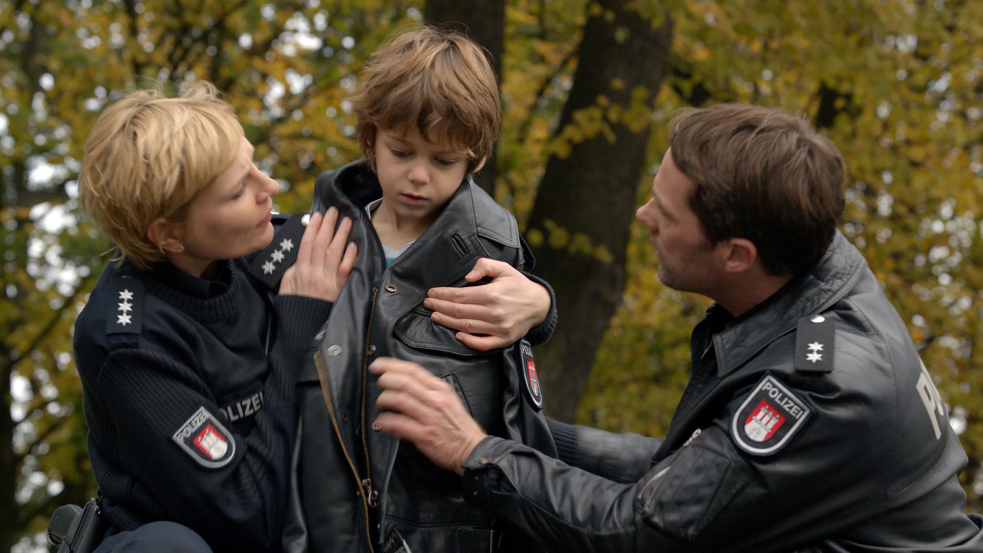 "Notruf Hafenkante - Das Buddelschiff": Die Polizisten Melanie Hansen (Sanna Englund) und Mattes Seeler (Matthias Schloo) knien neben dem kleinen Markus (Bruno Thiel). Melanie hat ihre Lederjacke um den Jungen gelegt. Sie hält ihn im Arm und sieht ihn besorgt an.