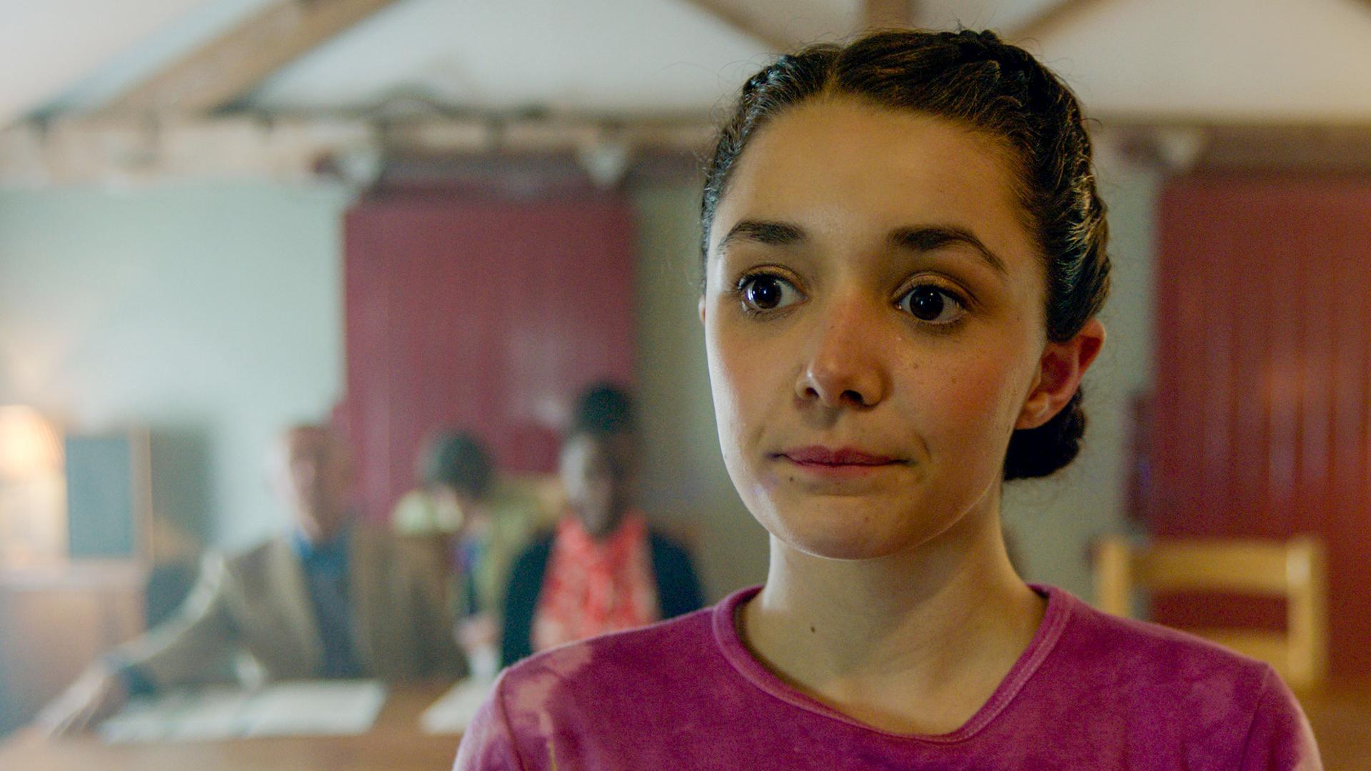 Addie steht im Vordergrund und schaut leicht an der Kamera vorbei. Im Hintergrund sitzen 3 Erwachsene an einem langen Holztisch mit Papieren vor sich liegend.