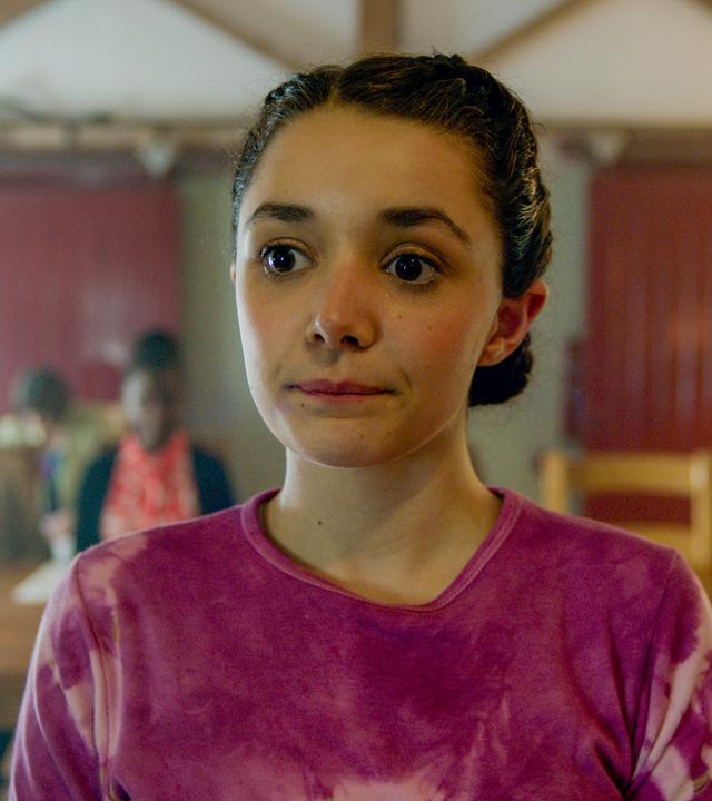 Addie steht im Vordergrund und schaut leicht an der Kamera vorbei. Im Hintergrund sitzen 3 Erwachsene an einem langen Holztisch mit Papieren vor sich liegend.