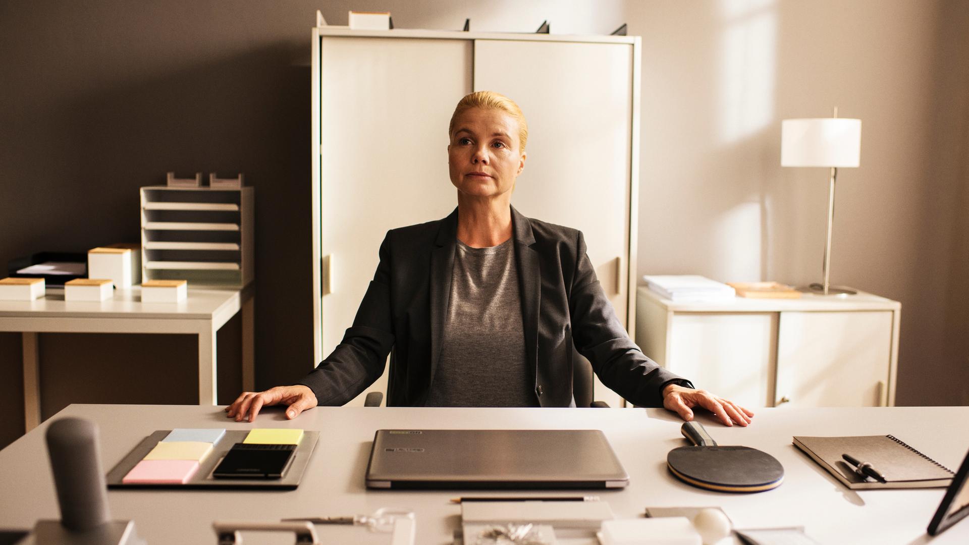 "Ella Schön - Das Ding mit der Liebe":Ella (Annette Frier) sitzt an einem sehr ordentlichen Schreibtisch in einem Büro und hat beide Hände auf den Tisch gelegt. Vor ihr auf dem Tisch befinden sich Schreibwaren, ein Laptop, ein Handy und ein Tischtennisschläger, alles wohlgeordnet.