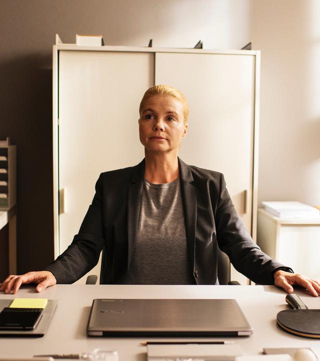 "Ella Schön - Das Ding mit der Liebe":Ella (Annette Frier) sitzt an einem sehr ordentlichen Schreibtisch in einem Büro und hat beide Hände auf den Tisch gelegt. Vor ihr auf dem Tisch befinden sich Schreibwaren, ein Laptop, ein Handy und ein Tischtennisschläger, alles wohlgeordnet.