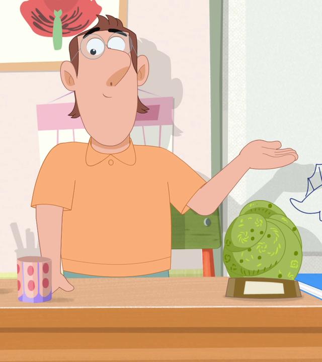 Herr Rebaud, Rebeccas Lehrer, steht hinter seinem Pult im Klassenzimmer und präsentiert das Dinosaurier-Fossil, welches auf dem Pult steht.