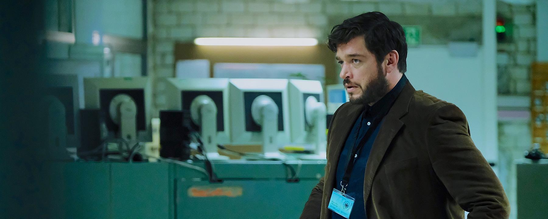 Martin Behrens (Ronald Zehrfeld) trägt ein dunkelbraunes Jackett und ein Schlüsselband mit Ausweis um den Hals. Im Hintergrund sieht man die Rückseiten mehrerer PC Bildschirme.