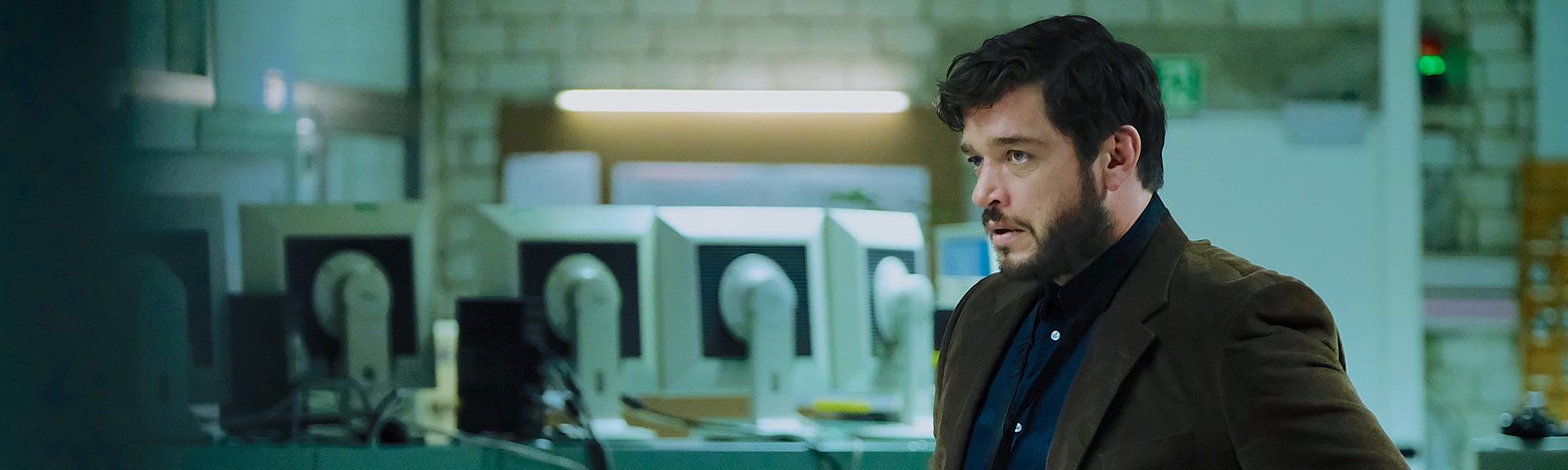 Martin Behrens (Ronald Zehrfeld) trägt ein dunkelbraunes Jackett und ein Schlüsselband mit Ausweis um den Hals. Im Hintergrund sieht man die Rückseiten mehrerer PC Bildschirme.