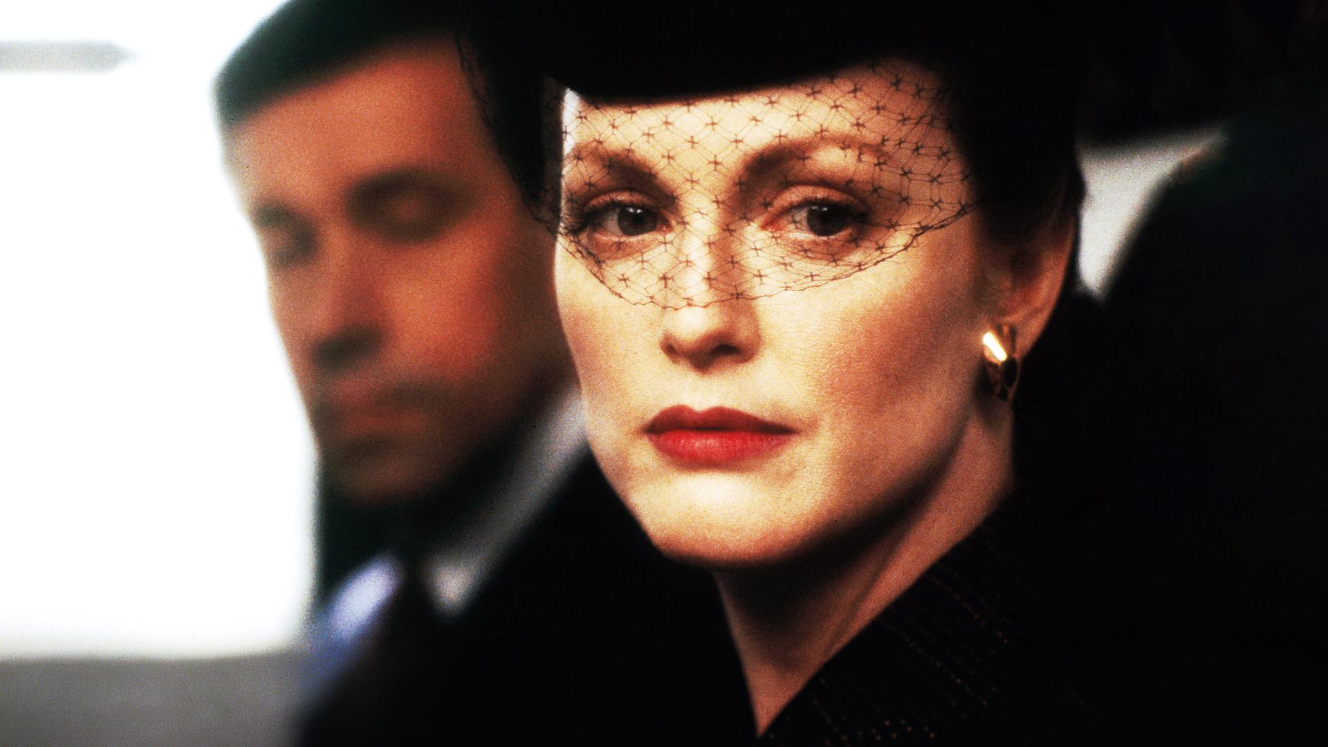 Sarah (Julianne Moore) mit einem schwarzen Gesichtsschleier aus Spitze vor ihrem Gesicht. Henry (Stephen Rea) ist etwas verschwommen im Hintergrund zu sehen.