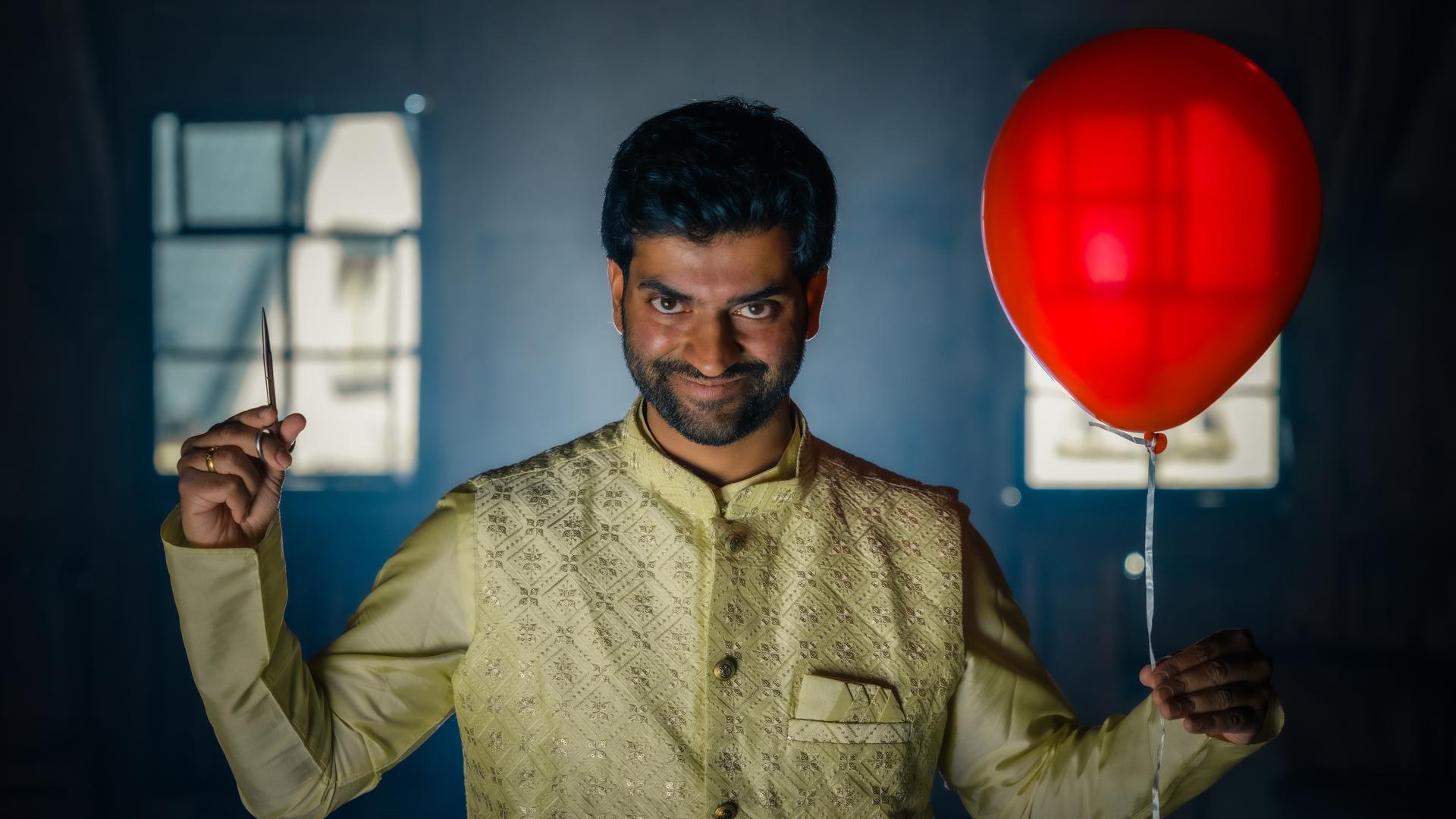 Der Hinduist Saran hält in der linken Hand einen roten Ballon und in der rechten Hand eine Schere. Dabei grinst er.