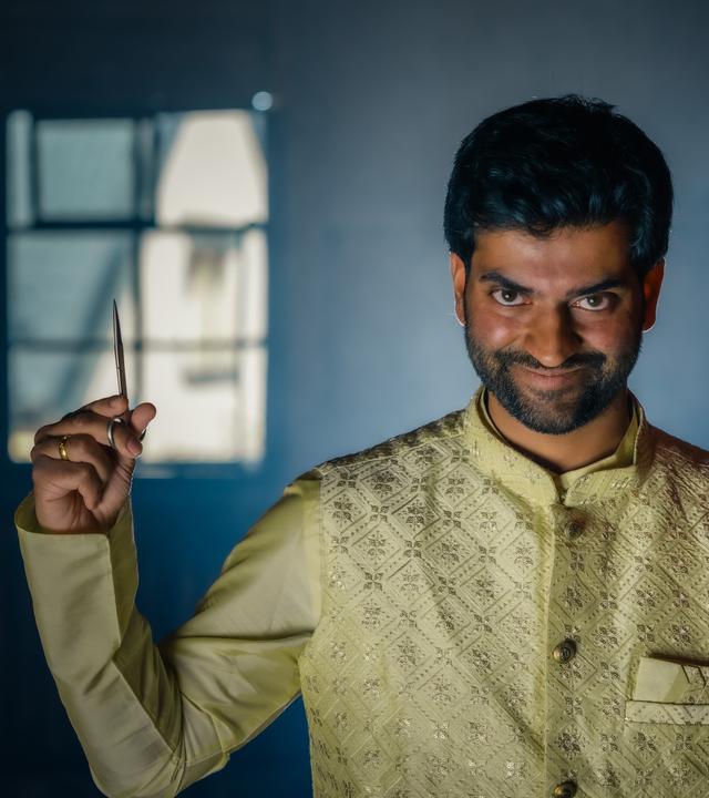 Der Hinduist Saran hält in der linken Hand einen roten Ballon und in der rechten Hand eine Schere. Dabei grinst er.