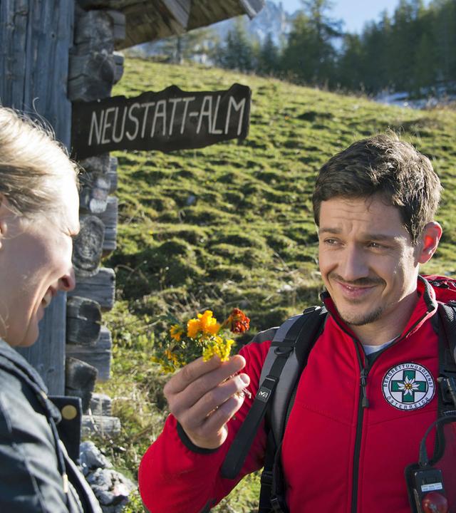"Die Bergretter - Das fremde Mädchen (2)": Vor der "Neustatt-Alm" stehen sich Emilie (Stefanie von Poser) und Tobias (Michael Brandl) gegenüber. Er hält einen kleinen selbst gepflückten Blumenstrauß in der Hand und lächelt sie an.