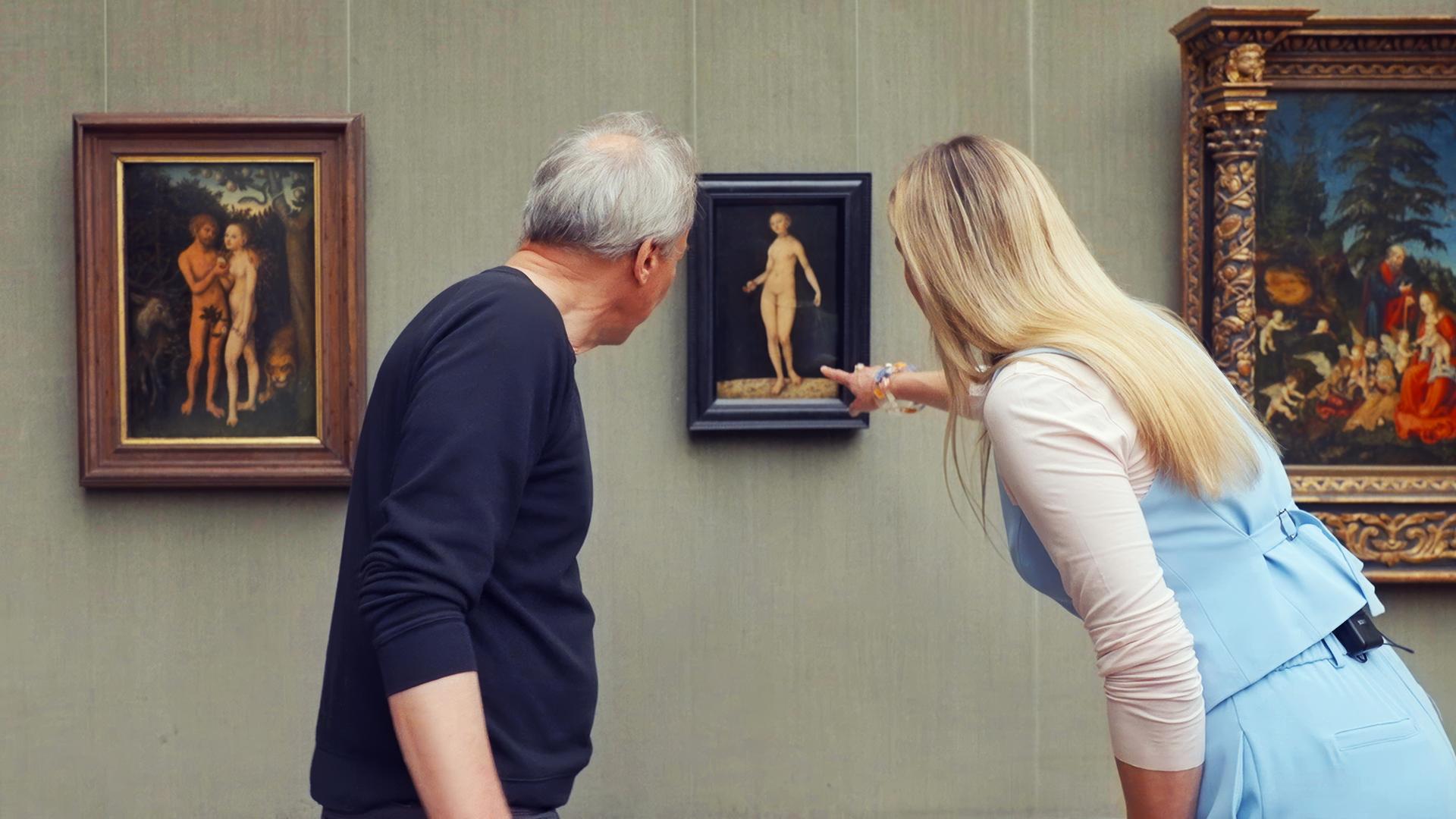 Ein älterer Mann und eine Frau betrachten ein Gemälde von Lucas Cranach, das Adam und Eva zeigt, in einer Gemäldegalerie.