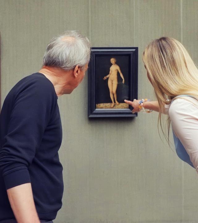 Ein älterer Mann und eine Frau betrachten ein Gemälde von Lucas Cranach, das Adam und Eva zeigt, in einer Gemäldegalerie.
