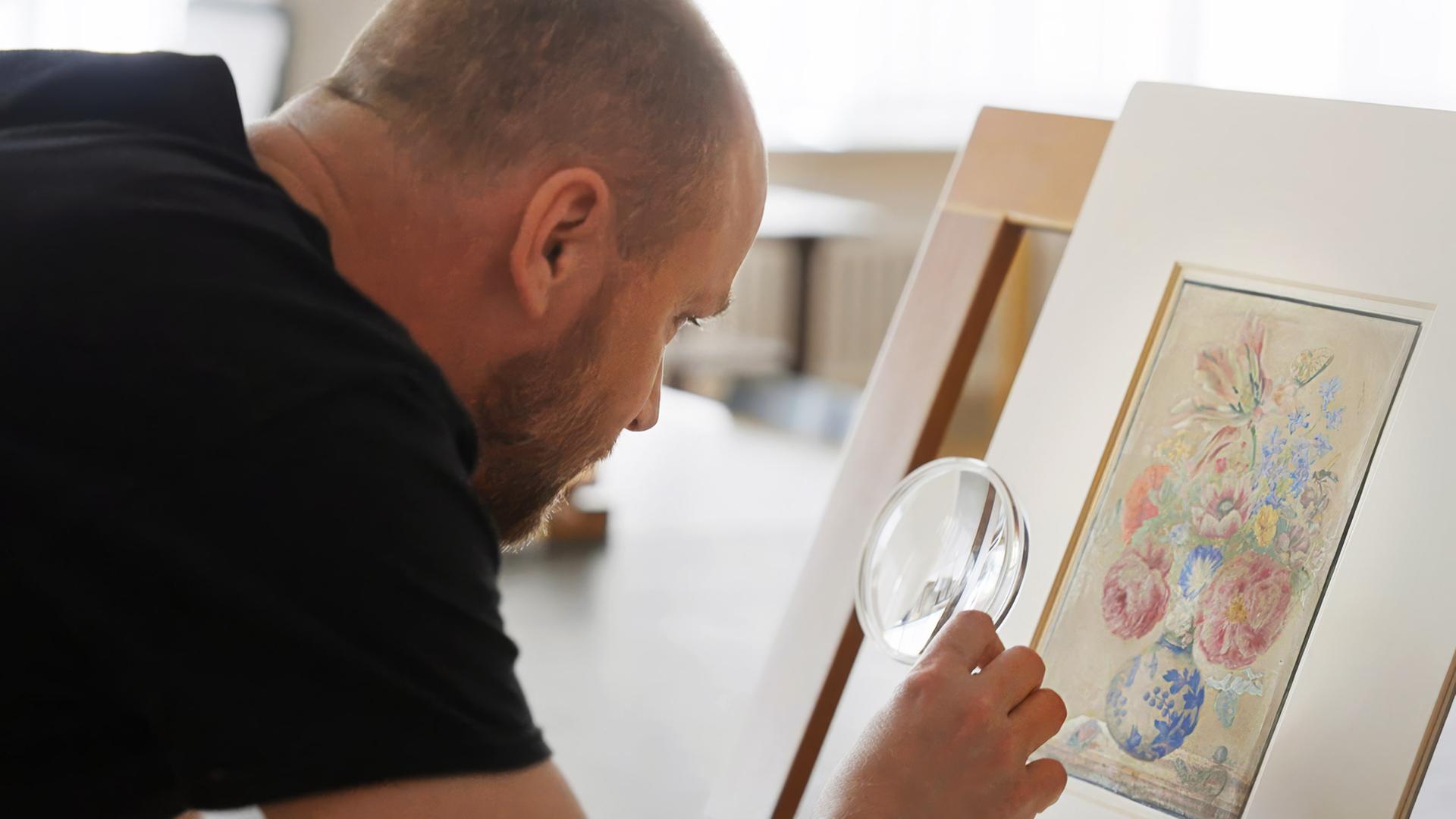 Ein Mann untersucht mit einer Lupe ein botanisches Aquarell mit Blumen von Maria Sibylla Merian.