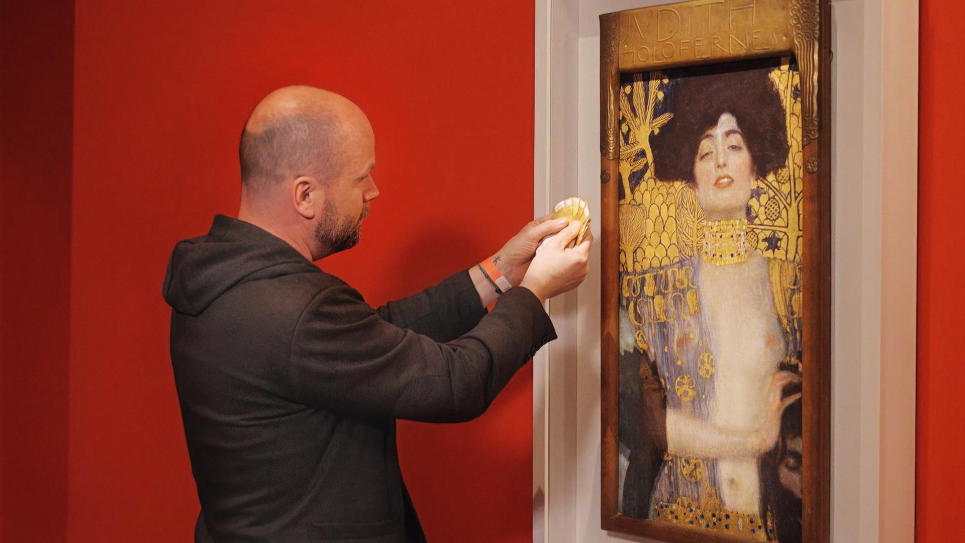 Ein Mann hält Farbmusterkarten an das Gemälde Judith und Holofernes von Gustav Klimt, das in einem weißen Rahmen vor einer roten Wand hängt.