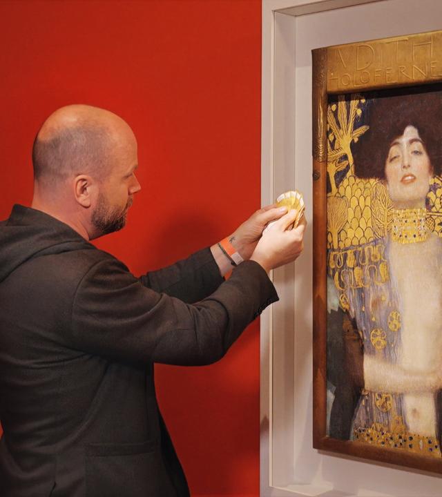 Ein Mann hält Farbmusterkarten an das Gemälde Judith und Holofernes von Gustav Klimt, das in einem weißen Rahmen vor einer roten Wand hängt.