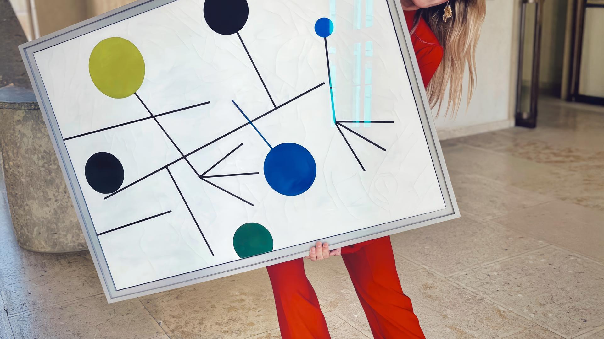 Frau in rotem Anzug posiert lachend mit einem großformatigen abstrakten Gemälde in einem hellen Museumsflur.