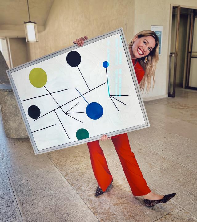 Frau in rotem Anzug posiert lachend mit einem großformatigen abstrakten Gemälde in einem hellen Museumsflur.