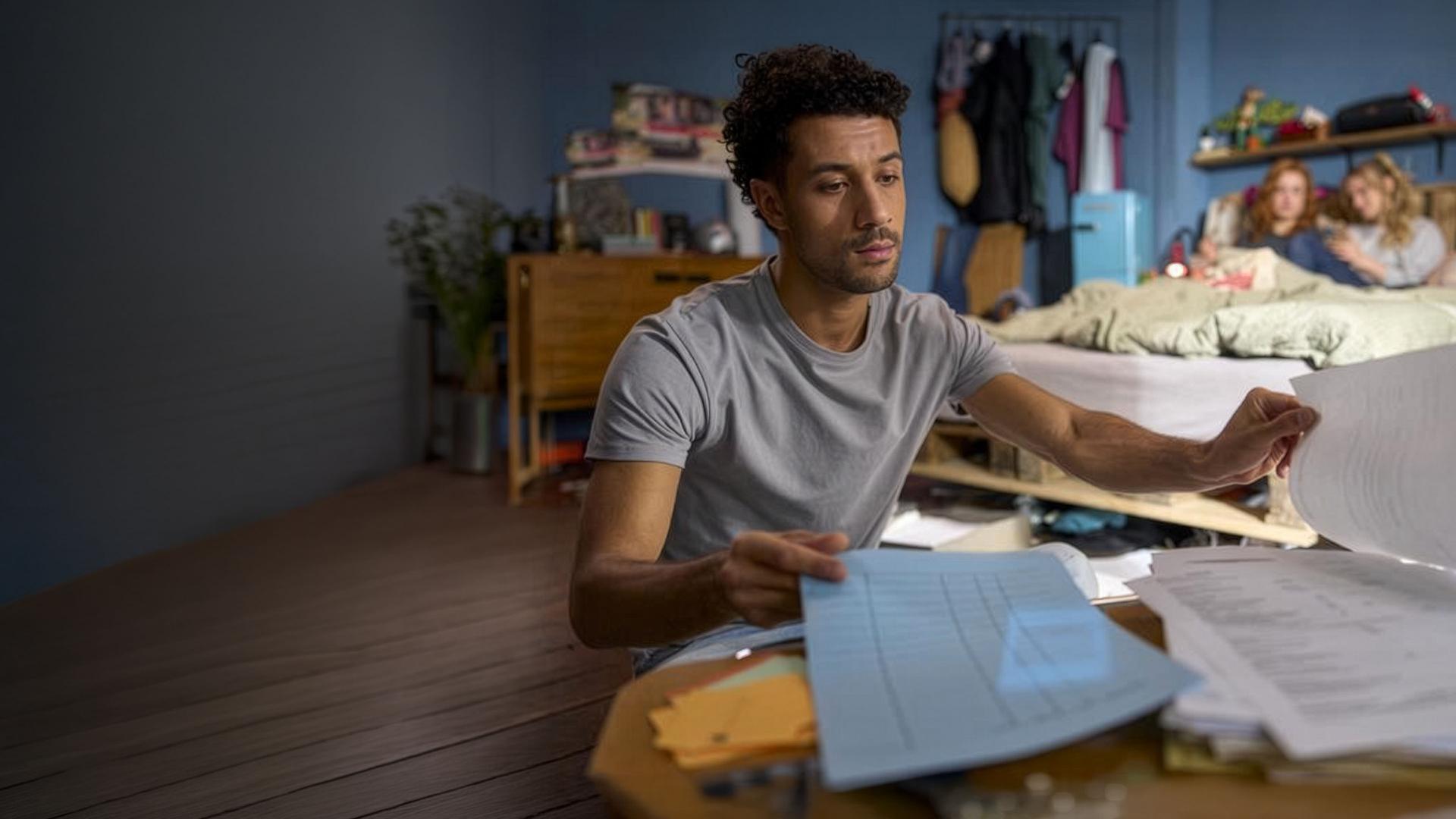 "Ich dich auch! - Das Gewitter": In einem Wohnraum: Yannik (Rojan Juan Barani) sitzt konzentriert vor einem Haufen Papiere. Caro (Meriel Hinsching) und Svenja (Leonie Wesselow) chillen auf dem Bett im Hintergrund.