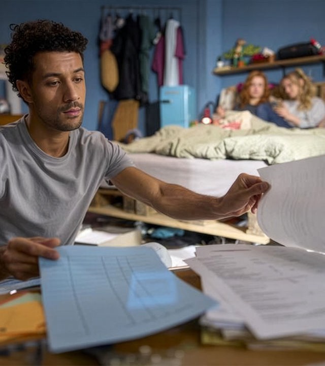 "Ich dich auch! - Das Gewitter": In einem Wohnraum: Yannik (Rojan Juan Barani) sitzt konzentriert vor einem Haufen Papiere. Caro (Meriel Hinsching) und Svenja (Leonie Wesselow) chillen auf dem Bett im Hintergrund.
