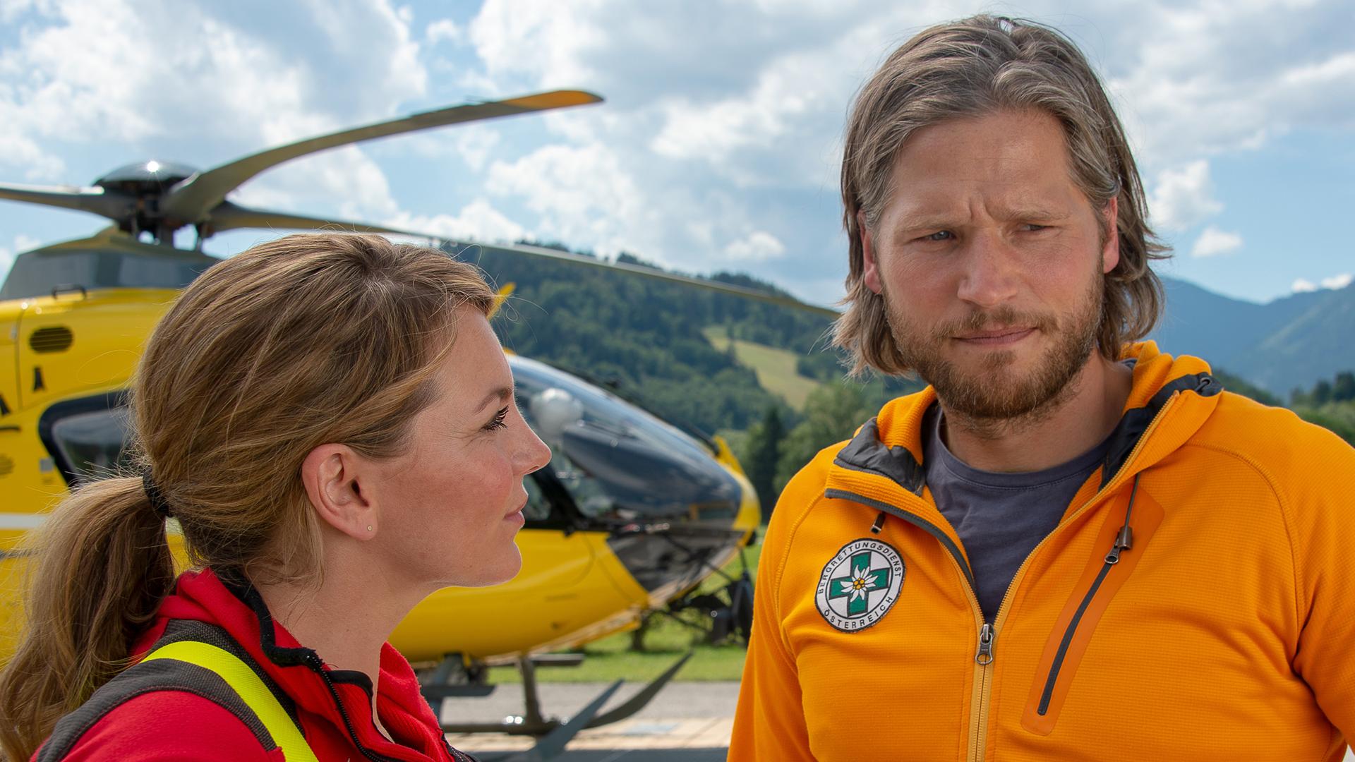 Markus und Katharina stehen nebeneinander vor dem Rettungshubschrauber. Katharina blickt Markus lächelnd an, der nachdenklich nach vorne schaut.