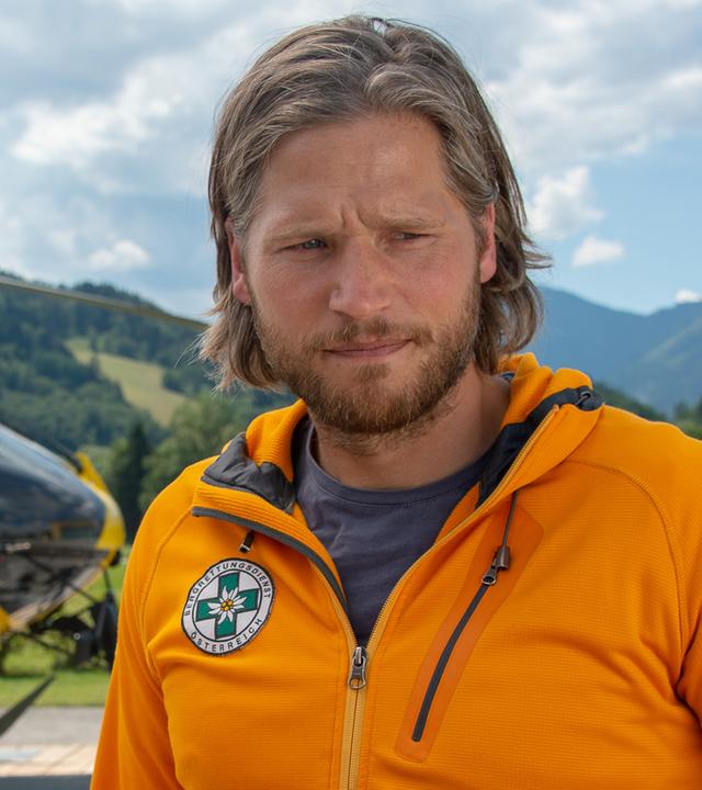 Markus und Katharina stehen nebeneinander vor dem Rettungshubschrauber. Katharina blickt Markus lächelnd an, der nachdenklich nach vorne schaut.