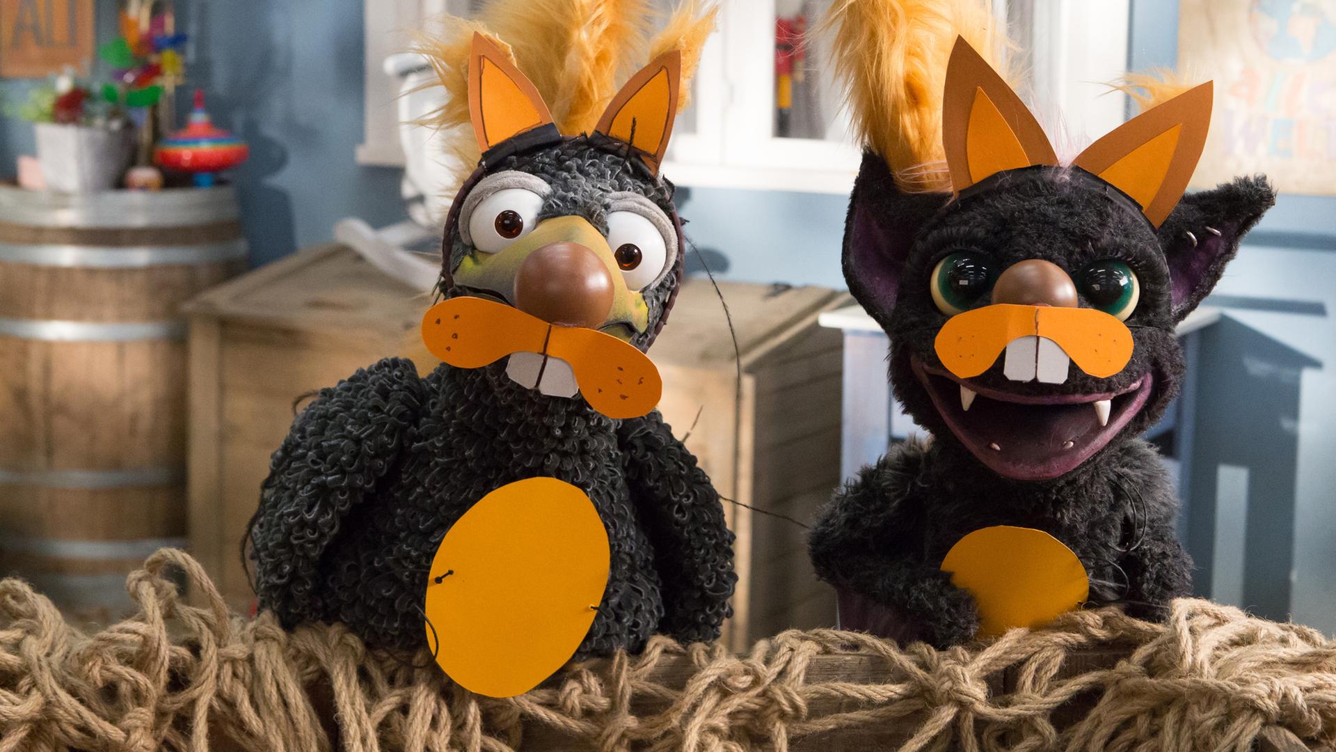 Rudi und Vampi hocken auf dem Hof vor dem Laden. Sie sind mit orangefarbenen Ohren, Schnauzen und Puschelschwänzen als Eichhörnchen verkleidet und rufen erwartungsvoll. 