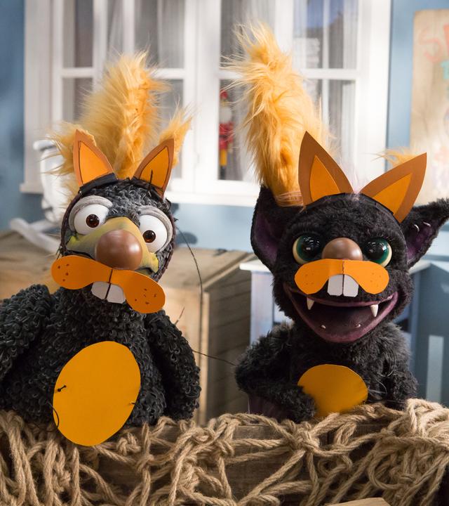 Rudi und Vampi hocken auf dem Hof vor dem Laden. Sie sind mit orangefarbenen Ohren, Schnauzen und Puschelschwänzen als Eichhörnchen verkleidet und rufen erwartungsvoll. 