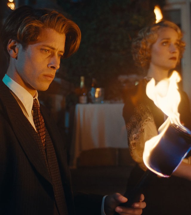 "This Is Not a Murder Mystery - Das Hummer-Telefon": René Magritte (Pierre Gervais) und Lee Miller (Florence Hall) stehen nebeneinander im Dunkeln. Im Vordergrund brennt eine Fackel, hinter den beiden sind weitere Lichter erkennbar.